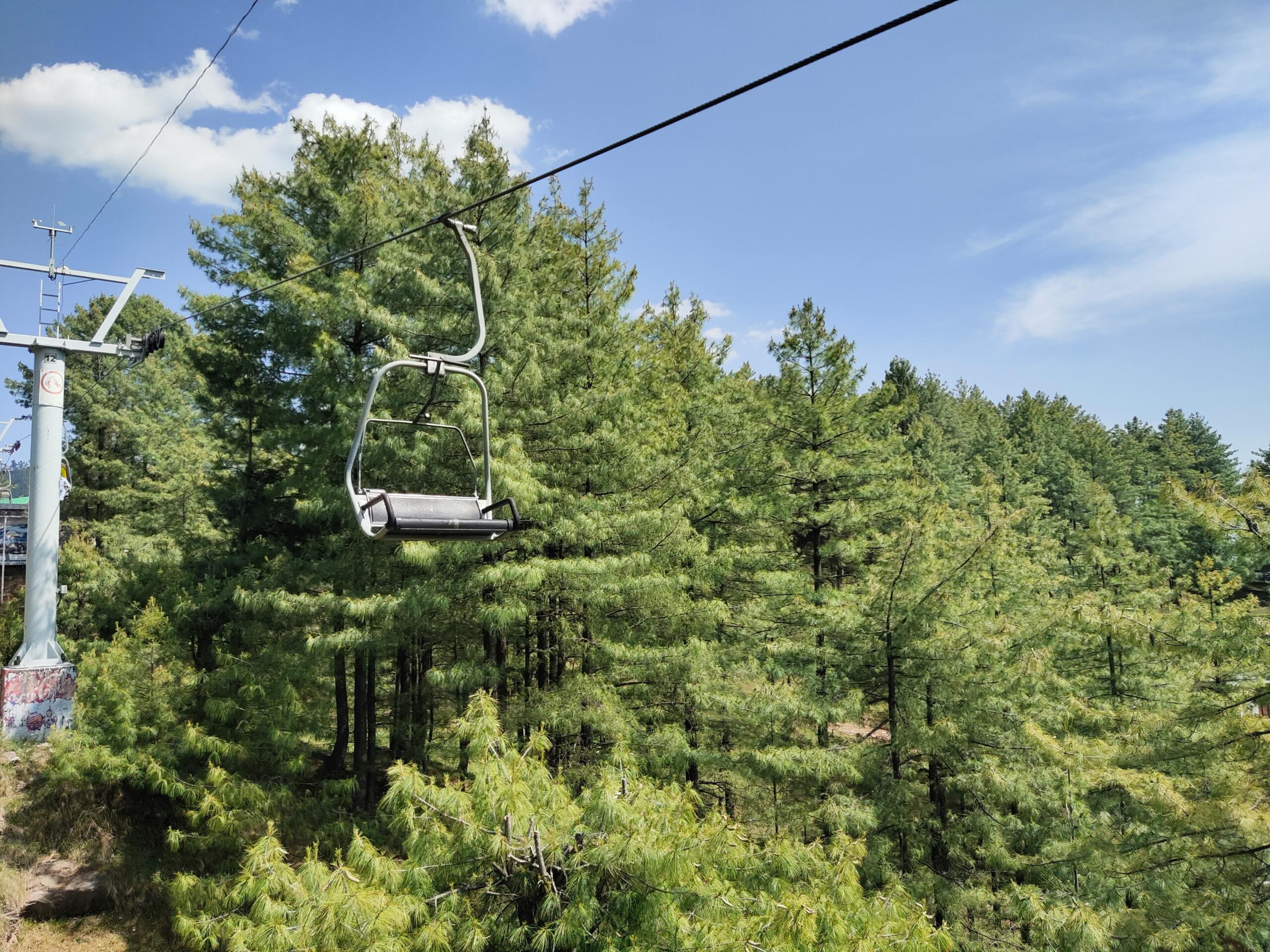 A scenic view of a chairlift over an evergreen forest in Murree, Pakistan on a sunny day.