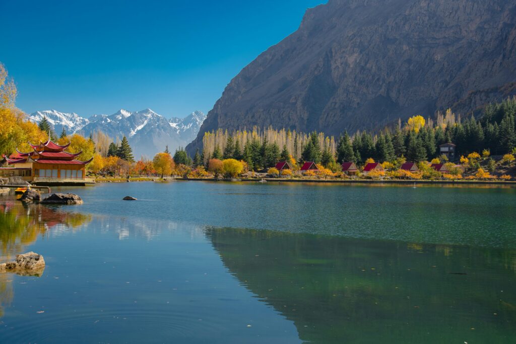 Peaceful autumn scene of a lake and mountains in Skardu, with vibrant foliage and a clear sky.