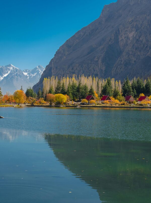 Peaceful autumn scene of a lake and mountains in Skardu, with vibrant foliage and a clear sky.